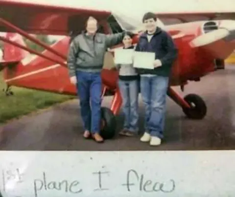 Two people holding certificates in front of a red airplane, with a handwritten note below
