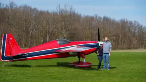 A man standing beside a red aerobatic airplane on the grass