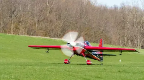 A red aerobatic airplane taxiing on the grass
