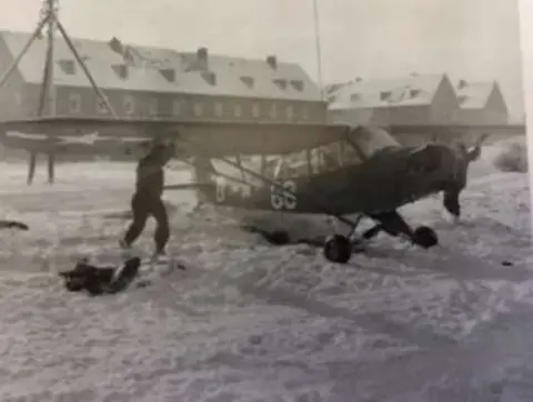 A Piper Cub on snow beside a large building, in an old photo