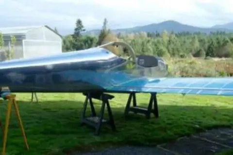An unpainted metal airplane on sawhorses outdoors with mountains behind