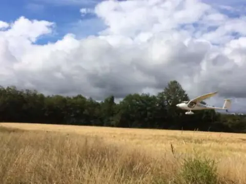 A white motorglider flying low over a dry grass field with trees behind