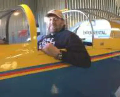 A man in a cap leaning on the cockpit edge of a yellow airplane in a hangar