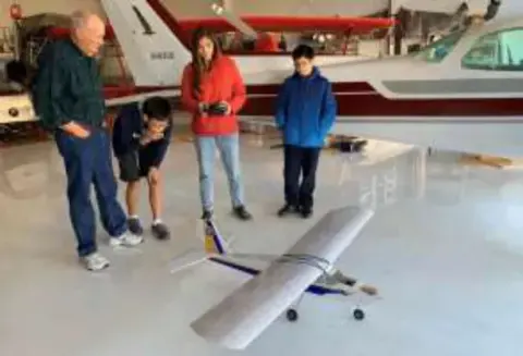 Students with an adult holding a finished model airplane in a hangar