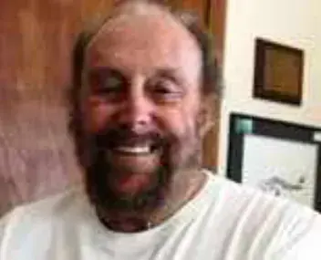 A smiling bald man with a white beard in a white T-shirt indoors