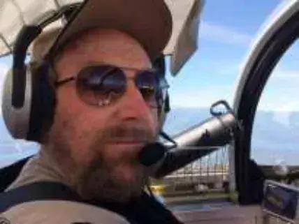 A smiling bearded pilot in a cap, sunglasses and headset