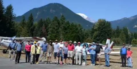 A large group of members posed in front of airplanes by a mountain