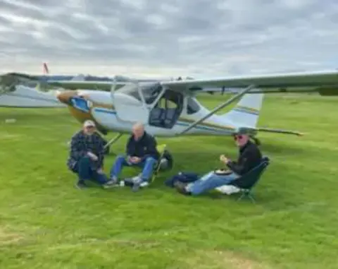 People sitting on the grass near parked light sport airplanes