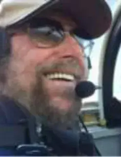 A smiling bearded pilot in a cap, sunglasses and headset