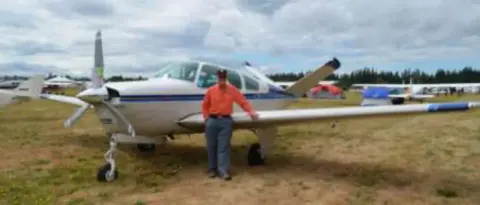 A man standing beside a white low-wing airplane on a grass field