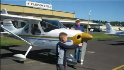 A boy and a man beside a white high-wing airplane on the ramp