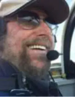 A smiling bearded pilot in a cap, sunglasses and headset