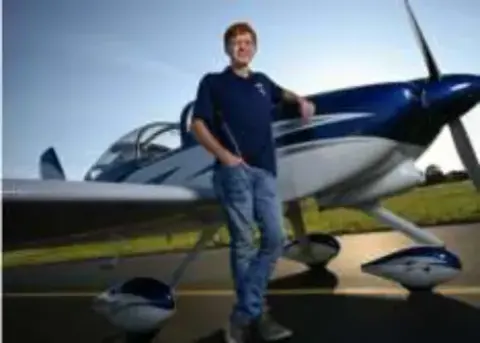A young man standing by the nose of a blue and white low-wing airplane