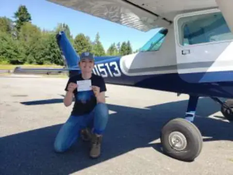 Madeline Patterson kneeling by the nose wheel of a high-wing airplane, holding her certificate