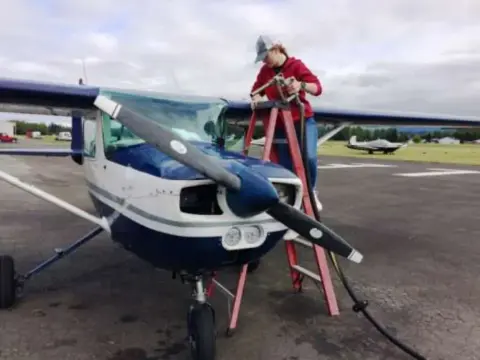 A person on a red stepladder working on the engine of a blue and white high-wing airplane