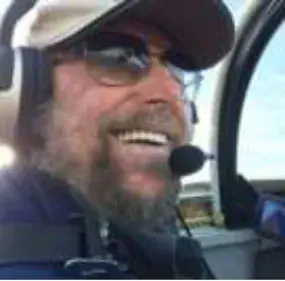 A smiling bearded pilot in a cap, sunglasses and headset