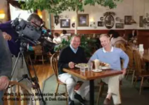 Two men seated at a restaurant table being filmed by a TV camera