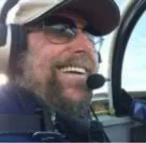 A smiling bearded pilot in a cap, sunglasses and headset