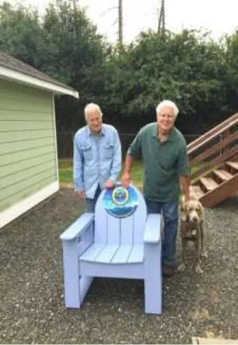 Two men with a painted Adirondack chair outside a shed