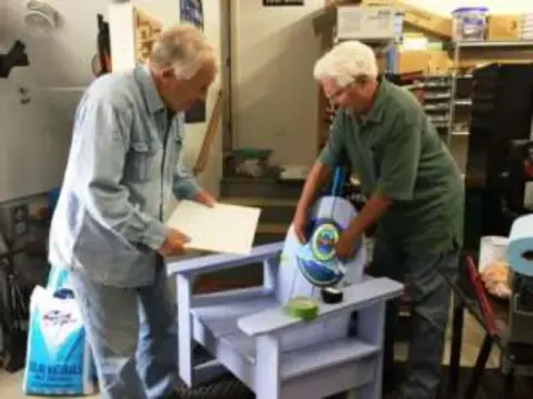 Two men painting an Adirondack chair in a workshop