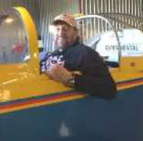 A man in a cap leaning on the cockpit edge of a yellow airplane in a hangar