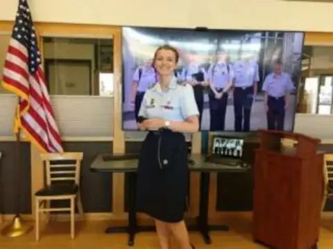 A woman giving a presentation in front of a screen with a flag beside her
