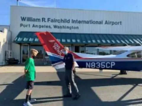 People near a red and white airplane in front of the Fairchild International Airport terminal