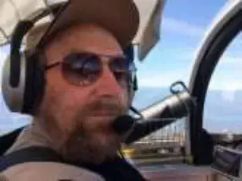 A bearded pilot in sunglasses and a headset in a cockpit