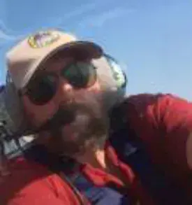 A bearded pilot in a cap, sunglasses and headset in a cockpit