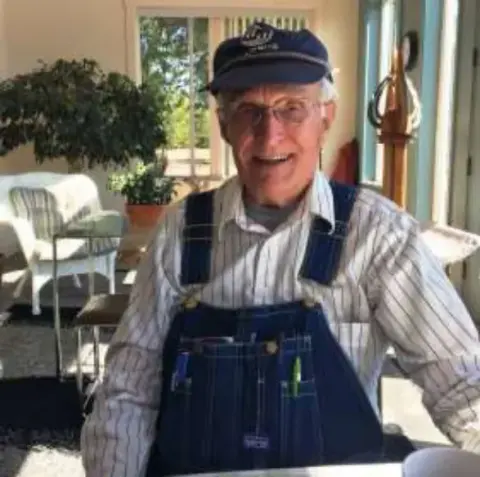 An older man in overalls and a cap smiling