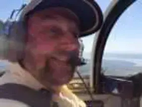 A bearded pilot in a headset and cap in a cockpit