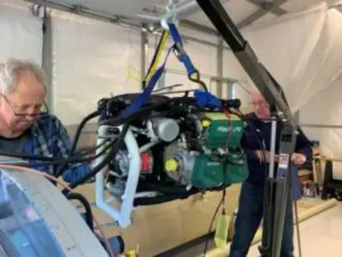 Two men using an engine hoist to mount an engine on an airplane in a hangar workshop