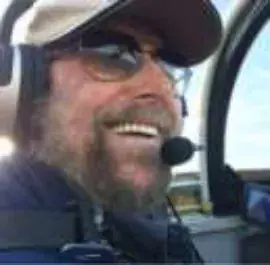 A smiling bearded pilot in a cap, sunglasses and headset in a cockpit
