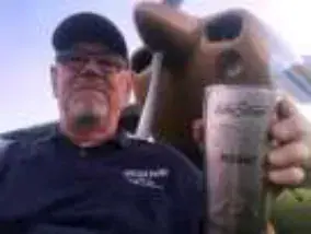 A man in a cap and dark shirt holding up a glass, a propeller behind him