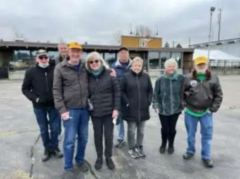A group of fly-out members standing on the ramp under cloudy skies