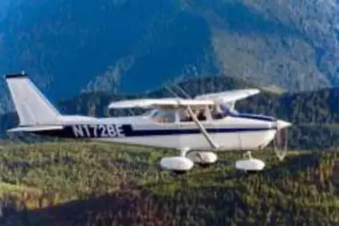 A white high-wing airplane parked on grass with mountains behind