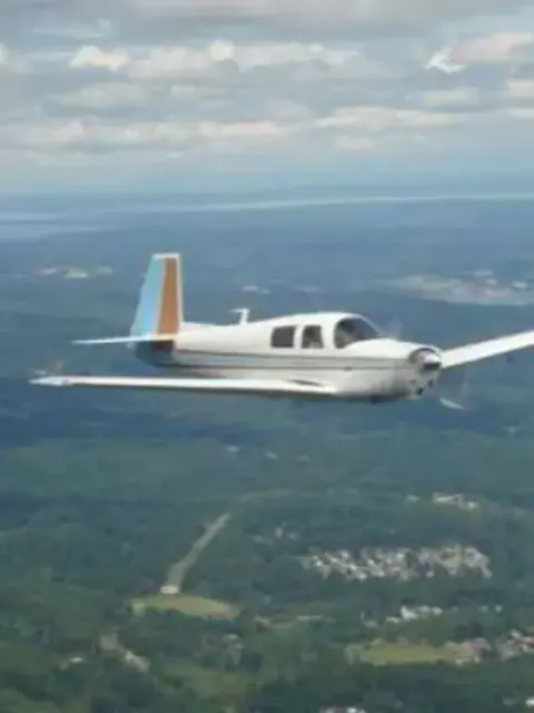 A white low-wing airplane in flight over forest