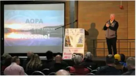 A speaker presenting to a seated audience with an AOPA slide on screen