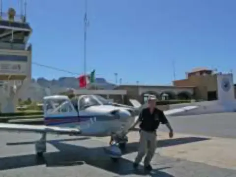 A man walking beside a low-wing airplane on an airport ramp, a control tower and a Mexican flag behind him