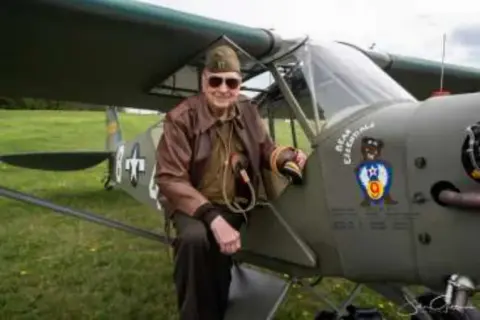 A man in a vintage flight jacket beside a liaison airplane with nose art