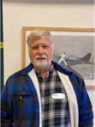 A bearded man in a plaid shirt standing in front of a framed airplane picture