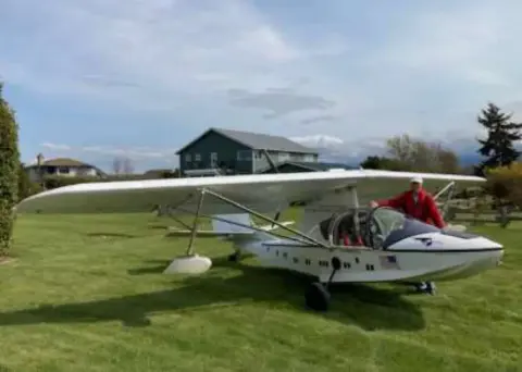 A man in the cockpit of a white amphibious airplane on grass