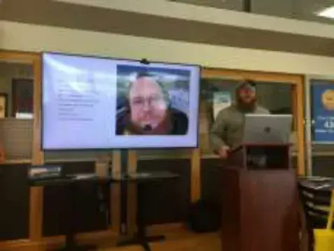 A man presenting next to a video call on a large screen
