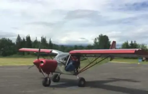 A red high-wing light airplane with its door open on the ramp