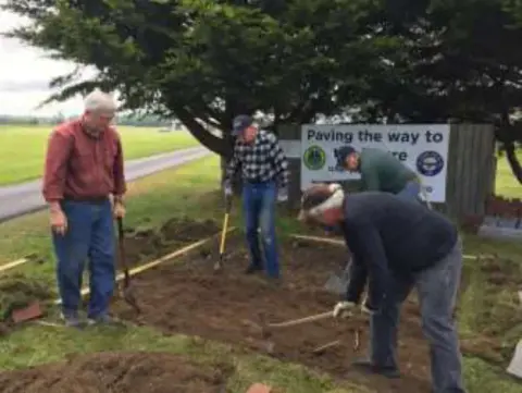 Volunteers digging and leveling ground beside a Paving the Way to the Future sign