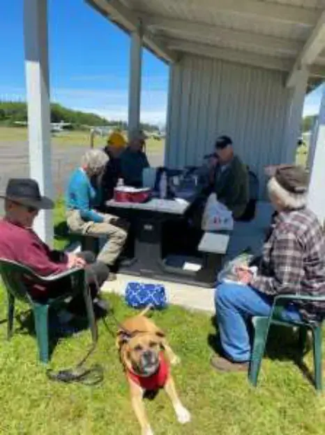 Members seated at picnic tables under a shelter with a dog nearby