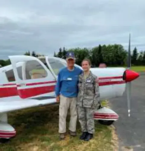 A man and a cadet standing by a red and white low-wing airplane