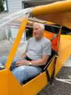 A white-haired man seated in the open cockpit of a yellow airplane