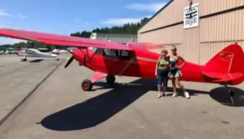 A red high-wing airplane on the ramp with people beside it