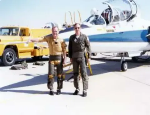 Two pilots in flight suits on the ramp beside a jet trainer